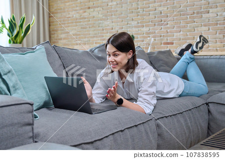 Surprised young woman looking at laptop screen lying on sofa at home Surprised young woman looking at laptop screen lying on sofa at home 107838595