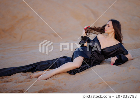 A woman in a black dress with a long train sits on a sand dune. A woman in a black dress with a long train sits on a sand dune. 107838788