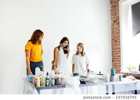Cute teenager in white T-shirt and two women in casual clothes are checking out acrylic-painted work. Young artist communicates with teachers in art workshop. Cute teenager in white T-shirt and two women in casual clothes are checking out acrylic-painted work. Young artist communicates with teachers in art workshop. 107838795