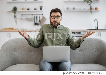 Confused male shrugging shoulders with laptop on couch Confused male shrugging shoulders with laptop on couch 107839330