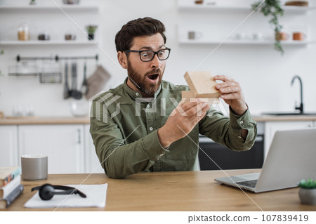 Man experiencing pleasant shock while unpacking parcel box Man experiencing pleasant shock while unpacking parcel box 107839419