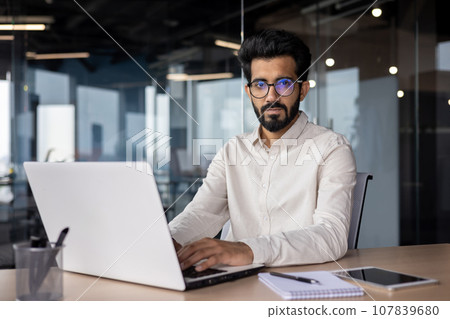 Portrait of an Indian man working in the office, sitting at the desk behind a laptop in a shirt and confidently looking at the camera. 107839680