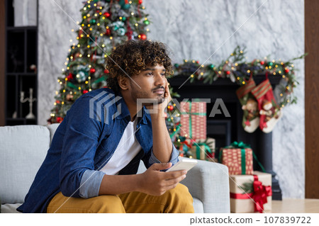 Bored man sitting on sofa for New Year holidays and Christmas, yawning hispanic man holding smartphone in living room near Christmas tree. Bored man sitting on sofa for New Year holidays and Christmas, yawning hispanic man holding smartphone in living room near Christmas tree. 107839722