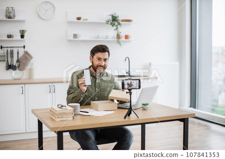 Young man recording video while unboxing smartphone indoors Young man recording video while unboxing smartphone indoors 107841353