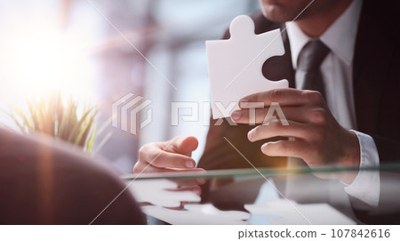 A business man sits at a table and holds a puzzle piece as a symbol of a business 107842616