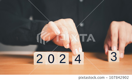 Male hand holding a wooden cube block with numbers 2024 on wood table. Goal in the new year 2024. Business development strategy, advancement and goal concept. 107842902