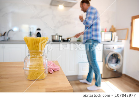 Focus on spaghetti in a glass container on a wooden table against the background of handsome man cooking dinner at home 107842977