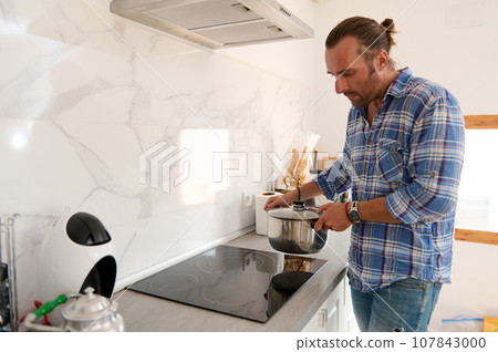 Portrait of a handsome young man putting a saucepan on the surface of an electric stove while cooking in home kitchen Portrait of a handsome young man putting a saucepan on the surface of an electric stove while cooking in home kitchen 107843000