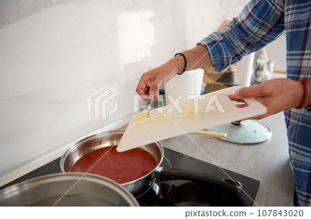Close-up of chef's hands putting chopped garlic from a cutting board into a saucepan with boiling tomato sauce Close-up of chef's hands putting chopped garlic from a cutting board into a saucepan with boiling tomato sauce 107843020