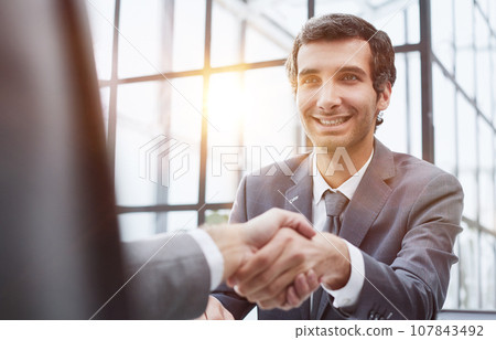 Portrait of confident mature specialist sitting at the table and reaching out his hand to the camera for a handshake Portrait of confident mature specialist sitting at the table and reaching out his hand to the camera for a handshake 107843492