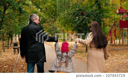 Loving parents hold hands of hopping little girl walking across autumn park. Parents with playful daughter enjoy fall season in city park. Happy mother and father play with little daughter in garden 107843729