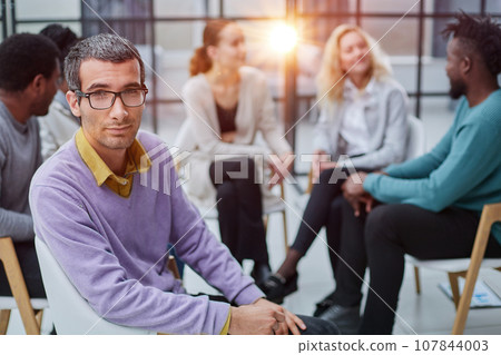 A happy young businessman, in a purple sweater, sits against the background of his colleagues 107844003