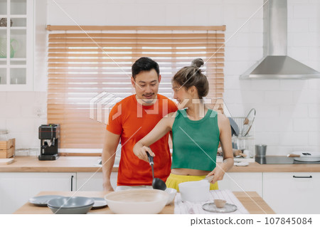 Husband looking at wife who cooking lunch for him. Happy couple in kitchen. Husband looking at wife who cooking lunch for him. Happy couple in kitchen. 107845084