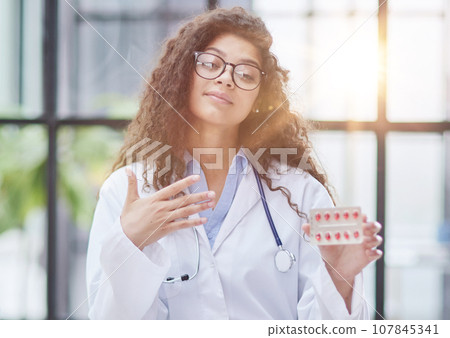 Young pharmacist woman holding pills while sitting at the table in the office Young pharmacist woman holding pills while sitting at the table in the office 107845341