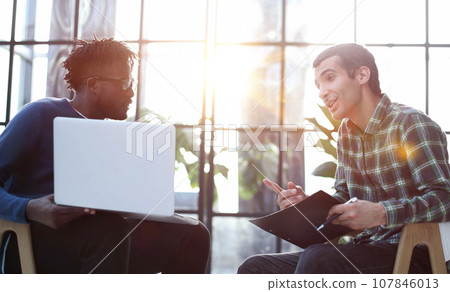 Friendly colleagues chat while sitting in the office, promising young manager shows a presentation on a laptop. Friendly colleagues chat while sitting in the office, promising young manager shows a presentation on a laptop. 107846013