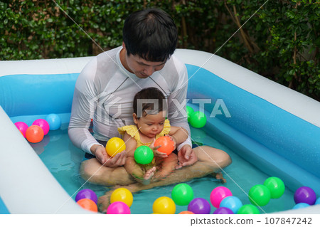 father and infant baby girl playing water with colorful plastic balls in inflatable pool 107847242