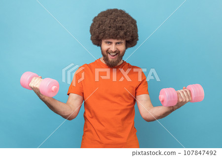 Portrait of strong man with Afro hairstyle wearing orange T-shirt raining biceps and triceps with pink dumbbells, screaming, showing his power. Indoor studio shot isolated on blue background. 107847492