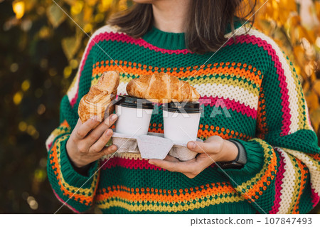 A lady in a colored striped sweater holding a coffee stand in front of yellow leaves 107847493