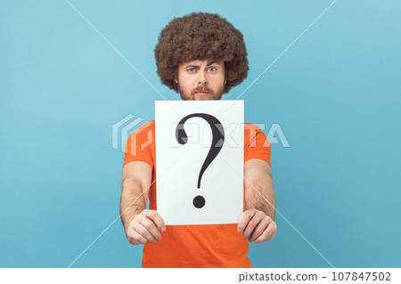 Portrait of man with Afro hairstyle wearing orange T-shirt holding question mark, finding smart solution, asking for advice, looking at camera. Indoor studio shot isolated on blue background. 107847502