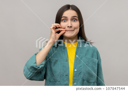 Portrait of attractive woman with mystery look making zip gesture to close mouth, keeping secret, zipping lips, wearing casual style jacket. Indoor studio shot isolated on gray background. Portrait of attractive woman with mystery look making zip gesture to close mouth, keeping secret, zipping lips, wearing casual style jacket. Indoor studio shot isolated on gray background. 107847544