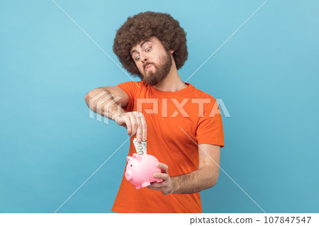 Portrait of man with Afro hairstyle wearing orange T-shirt investing, putting dollar banknote into piggy bank, proud with money savings. Indoor studio shot isolated on blue background. Portrait of man with Afro hairstyle wearing orange T-shirt investing, putting dollar banknote into piggy bank, proud with money savings. Indoor studio shot isolated on blue background. 107847547