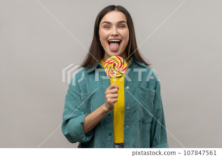 Childish attractive woman licking multicolor candy, wants to eat, looking at camera, showing tongue out, wearing casual style jacket. Indoor studio shot isolated on gray background. Childish attractive woman licking multicolor candy, wants to eat, looking at camera, showing tongue out, wearing casual style jacket. Indoor studio shot isolated on gray background. 107847561