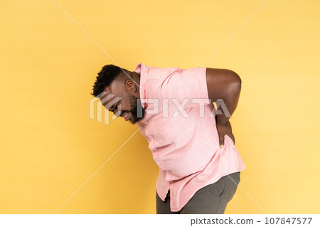Portrait of exhausted unhealthy man wearing pink shirt touching aching back, suffering lower lumbar discomfort, muscle pain, injured spine disk. Indoor studio shot isolated on yellow background. Portrait of exhausted unhealthy man wearing pink shirt touching aching back, suffering lower lumbar discomfort, muscle pain, injured spine disk. Indoor studio shot isolated on yellow background. 107847577