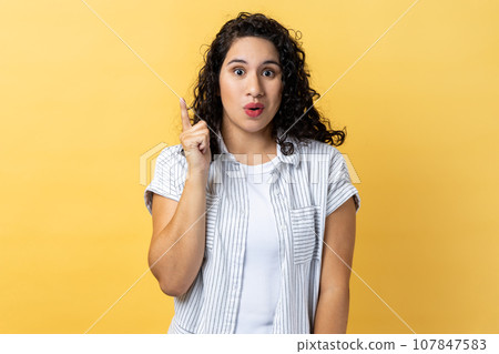 Portrait of excited amazed woman with dark wavy hair pointing upwards, looking at camera with open mouth, having new great idea. Indoor studio shot isolated on yellow background. Portrait of excited amazed woman with dark wavy hair pointing upwards, looking at camera with open mouth, having new great idea. Indoor studio shot isolated on yellow background. 107847583