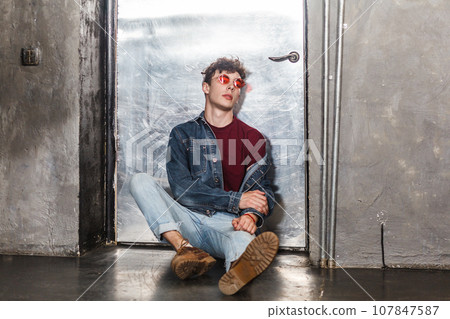 Full length portrait of calm attractive relaxed fashionable model man in bright red sunglasses and denim casual style, sitting near metallic door, looking at camera. Indoor studio shot. 107847587