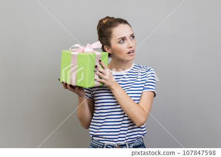 Portrait of woman wearing striped T-shirt shaking wrapped present box, being interested what inside, looking away with curious facial expression. Indoor studio shot isolated on gray background. 107847598