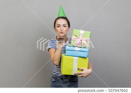 Portrait of flirting woman wearing striped T-shirt and party cone, holding stack of present boxes, sending air kiss. Indoor studio shot isolated on gray background. 107847602