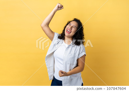 Portrait of excited happy woman with dark wavy hair expressing winning gesture with raised fists and screaming, celebrating victory. Indoor studio shot isolated on yellow background. Portrait of excited happy woman with dark wavy hair expressing winning gesture with raised fists and screaming, celebrating victory. Indoor studio shot isolated on yellow background. 107847604