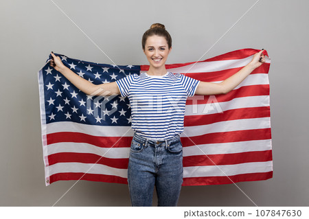 Portrait of joyful young adult smiling woman wearing striped T-shirt holding big american flag, expressing positive emotions and happiness. Indoor studio shot isolated on gray background. Portrait of joyful young adult smiling woman wearing striped T-shirt holding big american flag, expressing positive emotions and happiness. Indoor studio shot isolated on gray background. 107847630