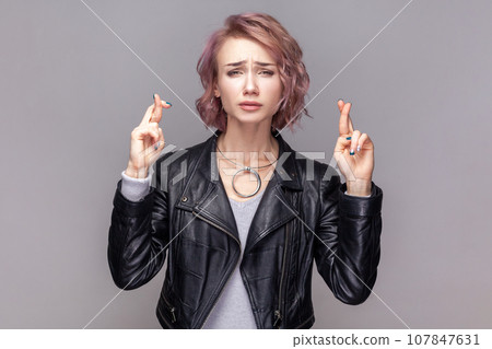 Portrait of winsome woman with short hairstyle standing with crossed fingers, making for good luck, looking at camera, wearing black leather jacket. Indoor studio shot isolated on grey background. Portrait of winsome woman with short hairstyle standing with crossed fingers, making for good luck, looking at camera, wearing black leather jacket. Indoor studio shot isolated on grey background. 107847631