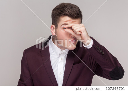 Portrait of attractive handsome young man keeps her finger near eyes, showing small gesture, peeping, wearing violet suit and white shirt. Indoor studio shot isolated on grey background. 107847701