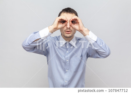 Portrait of curious funny young adult man standing hands near eyes, making binocular gesture, looking at something interesting, wearing light blue shirt. Indoor studio shot isolated on gray background 107847712
