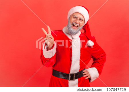 Amazed elderly man with gray beard wearing santa claus costume standing with raised hands and showing v sign or peace, victory gesture. Indoor studio shot isolated on red background. 107847729