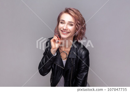 Portrait of charming beautiful adorable woman with short hairstyle standing looking at camera with toothy smile, wearing black leather jacket. Indoor studio shot isolated on grey background. Portrait of charming beautiful adorable woman with short hairstyle standing looking at camera with toothy smile, wearing black leather jacket. Indoor studio shot isolated on grey background. 107847735