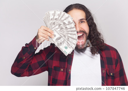 Portrait of amazed surprised excited bearded man with long curly hair in checkered red shirt covering half of face with dollar banknotes. Indoor studio shot isolated on gray background. Portrait of amazed surprised excited bearded man with long curly hair in checkered red shirt covering half of face with dollar banknotes. Indoor studio shot isolated on gray background. 107847746