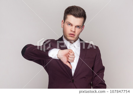 Portrait of handsome man shows disapproval sign, keeps thumb down, expresses dislike, frowns face in discontent, wearing violet suit and white shirt. Indoor studio shot isolated on grey background. 107847754
