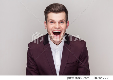 Portrait of handsome man laughs at something funny, looking at camera, having fun, expressing positive emotions, wearing violet suit and white shirt. Indoor studio shot isolated on grey background. 107847770
