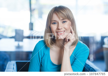 Portrait of smiling happy satisfied young woman with blonde hair in blue shirt sitting alone, looking at camera with toothy smile, being in good mood. Indoor shot in cafe with big window on background Portrait of smiling happy satisfied young woman with blonde hair in blue shirt sitting alone, looking at camera with toothy smile, being in good mood. Indoor shot in cafe with big window on background 107847785