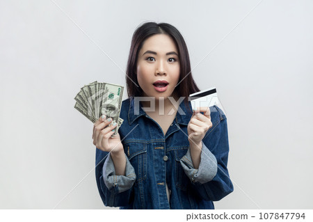 Portrait of shocked surprised brunette woman in blue denim jacket standing looking at camera with open mouth, holding dollar banknotes and credit card. Indoor studio shot isolated on gray background. 107847794
