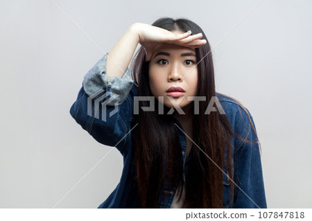 Portrait of woman in blue denim jacket standing keeping palm over head and looking attentively far away, peering with expectation at long distance. Indoor studio shot isolated on gray background. Portrait of woman in blue denim jacket standing keeping palm over head and looking attentively far away, peering with expectation at long distance. Indoor studio shot isolated on gray background. 107847818