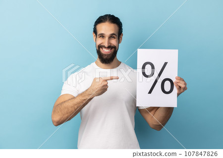 Portrait of attractive optimistic man with beard wearing white T-shirt holding and pointing paper with percent sign inscription, looking at camera. Indoor studio shot isolated on blue background. 107847826