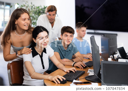 Girl stands behind sitting students, looks at monitor and helps in execution of programing teamwork Girl stands behind sitting students, looks at monitor and helps in execution of programing teamwork 107847834