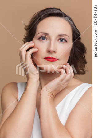 Closeup portrait of calm attractive beautiful middle aged woman with wavy hair and makeup, looking at camera, wearing white dress. Indoor studio shot isolated on light brown background. Closeup portrait of calm attractive beautiful middle aged woman with wavy hair and makeup, looking at camera, wearing white dress. Indoor studio shot isolated on light brown background. 107847835
