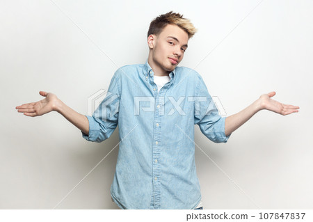 Portrait of doubtful puzzled attractive man wearing denim shirt standing with spread hands, looking at camera and shrugging shoulders. Indoor studio shot isolated on gray background. Portrait of doubtful puzzled attractive man wearing denim shirt standing with spread hands, looking at camera and shrugging shoulders. Indoor studio shot isolated on gray background. 107847837