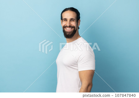 Side view of bearded handsome man wearing white T-shirt standing looking at camera with satisfied face and smiling, expressing happiness. Indoor studio shot isolated on blue background. Side view of bearded handsome man wearing white T-shirt standing looking at camera with satisfied face and smiling, expressing happiness. Indoor studio shot isolated on blue background. 107847840