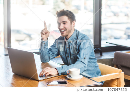 Portrait of excited overjoyed young man working on laptop and raised finger up, having good idea, has online meeting, discussing project. Indoor shot near big window, cafe background. 107847853